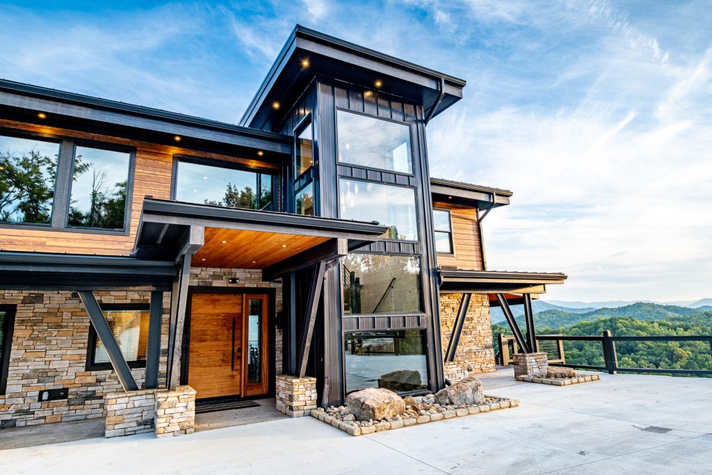 Modern timber-and-stone cabin entrance with mountain views beyond the driveway.
